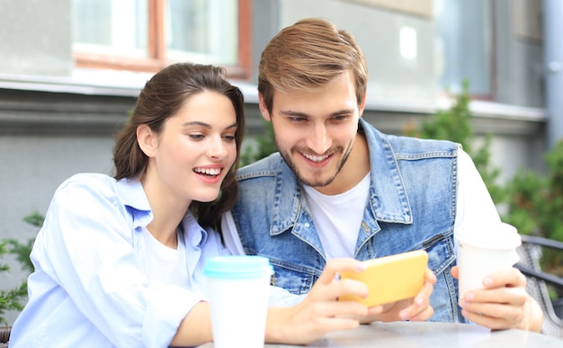 Aantrekkelijke jonge verliefde paar drinken koffie terwijl ze buiten aan de cafétafel zitten, met behulp van mobiele telefoon.