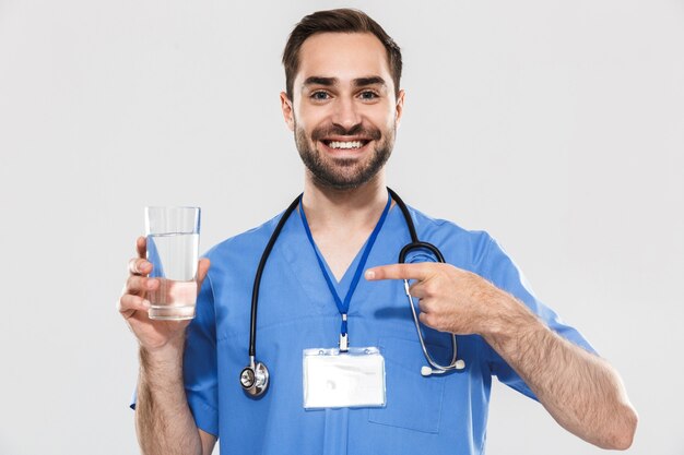 Aantrekkelijke jonge, vrolijke mannelijke arts met een unifrom die over een witte muur staat en een glas water toont