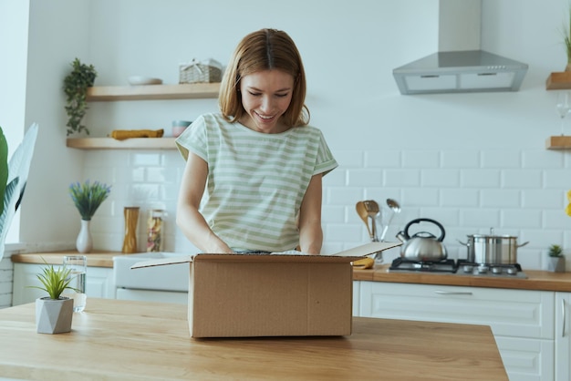 Aantrekkelijke jonge vrouw die de doos uitpakt terwijl ze in de huishoudelijke keuken staat