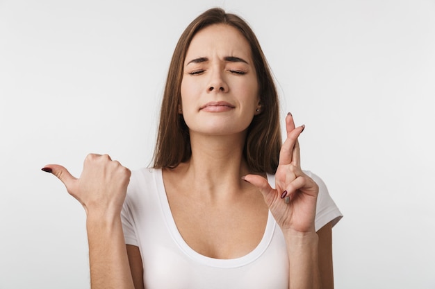 Aantrekkelijke jonge vrouw die geïsoleerd over een witte muur staat, met de vingers gekruist voor geluk