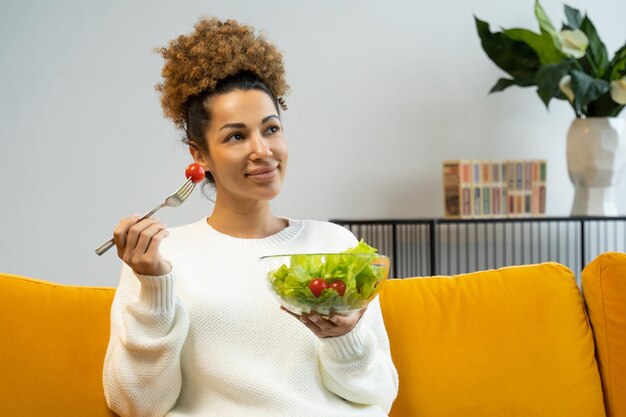 Aantrekkelijke jonge vrouw die groentesalade eet van een bord dat thuis op de bank zit