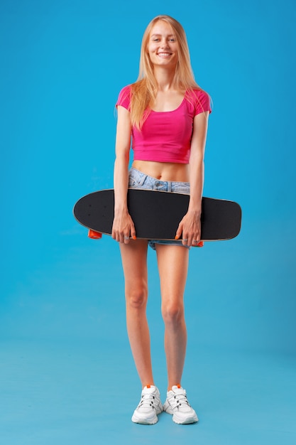 Aantrekkelijke jonge vrouw die haar skateboard over blauwe achtergrond houdt