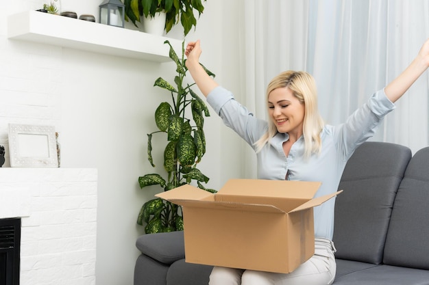 Aantrekkelijke jonge vrouw die hartelijk lacht terwijl ze een kartonnen doos met verpakking in haar handen houdt in haar woonkamer