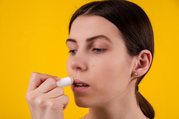 Aantrekkelijke jonge vrouw die hygiënische lippenstift op gele achtergrond gebruikt. lippen zorg en bescherming.