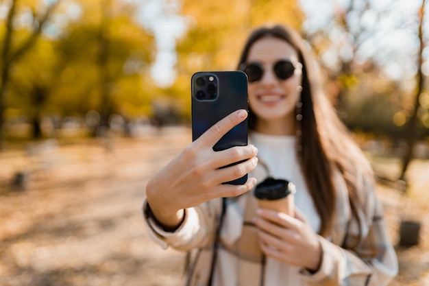 Aantrekkelijke jonge vrouw die in de herfst loopt en een jas draagt met behulp van de telefoon