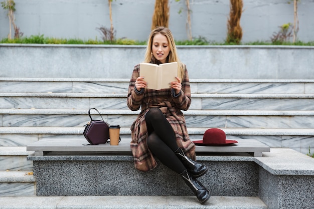 Foto aantrekkelijke jonge vrouw die jas draagt die in openlucht zit, een boek leest