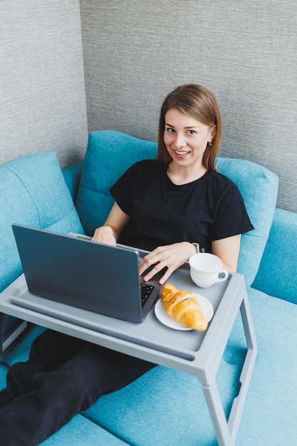 Aantrekkelijke jonge vrouw die laptopcomputer gebruikt terwijl ze thuis op de bank zit met koffie en croissants aan het werk Online werken op afstand