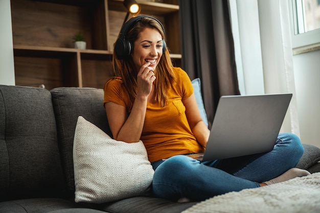 Aantrekkelijke jonge vrouw die met gekruiste benen op de bank zit en haar laptop en koptelefoon gebruikt om een videochat te maken met iemand thuis.