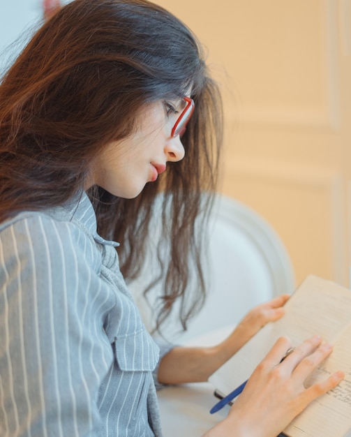 Aantrekkelijke jonge vrouw die met pen in hand op plannen denken en lijst schrijven om in blocnote te doen die van recreatietijd in modieuze koffie genieten