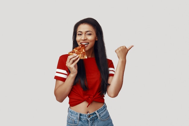 Aantrekkelijke jonge vrouw die pizza eet en naar de camera kijkt terwijl ze tegen een grijze achtergrond staat