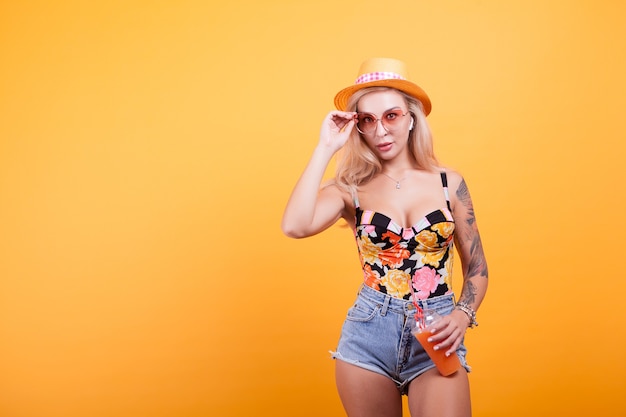 Aantrekkelijke jonge vrouw die sinaasappelsap drinkt over gele achtergrond met zonnebril in studio