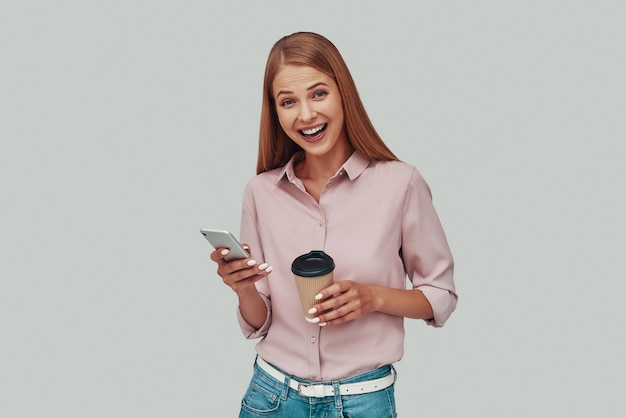 Aantrekkelijke jonge vrouw die slimme telefoon gebruikt en glimlacht terwijl ze tegen een grijze achtergrond staat