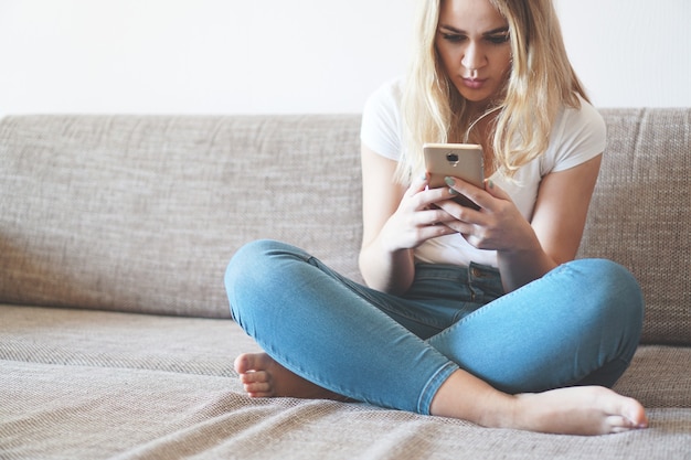 Aantrekkelijke jonge vrouw die thuis op een bank ontspant en een smartphone met touchscreen gebruikt om vanuit huis te socializen en te netwerken