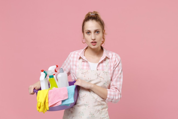 Aantrekkelijke jonge vrouw huisvrouw in schort houden bekken met wasmiddelflessen wassen reinigingsmiddelen terwijl het doen van huishoudelijk werk geïsoleerd op roze achtergrond studio portret. Huishoudelijk concept. Camera kijken.