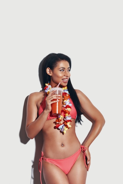 Aantrekkelijke jonge vrouw in bikini die verfrissende cocktail drinkt en glimlacht terwijl ze tegen een grijze achtergrond staat