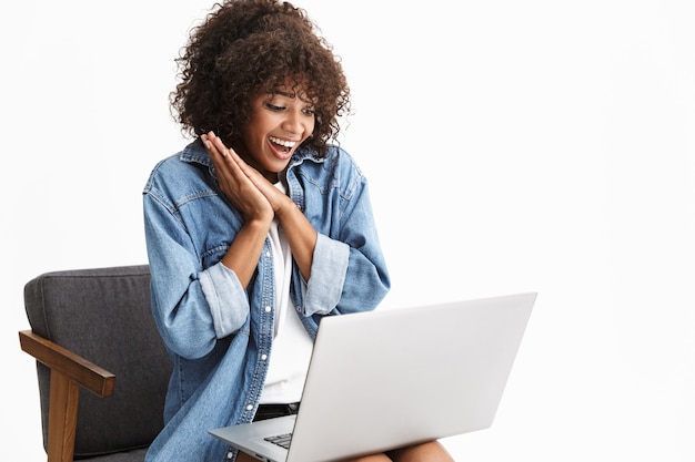 Aantrekkelijke jonge vrouw in denim zittend in een stoel geïsoleerd over een witte muur, werkend op laptopcomputer, chattend