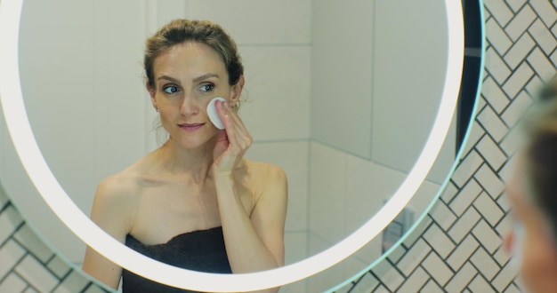 Aantrekkelijke jonge vrouw in een handdoek die het gezicht schoonmaakt met een wattenschijfje kijken naar spiegel make-up verwijderen Schoonheidsbehandeling huidverzorging concept close-up weergave