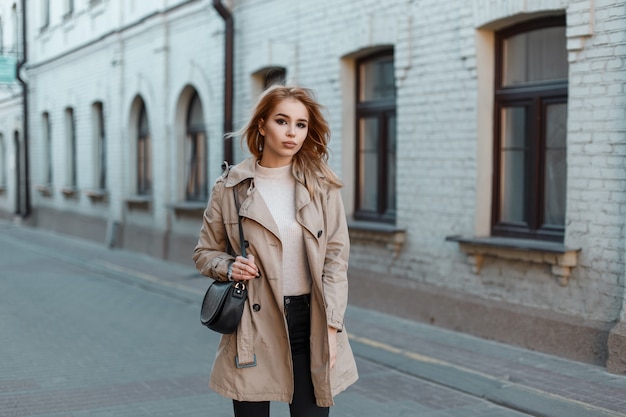Aantrekkelijke jonge vrouw in een stijlvolle lichte jas in een wit T-shirt in spijkerbroek met een zwart lederen tas op straat in de buurt van een wit bakstenen gebouw. Stijlvol blond meisje loopt door de stad.