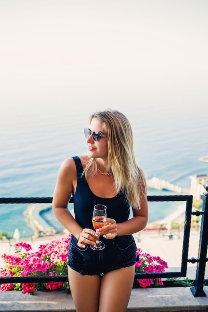 Aantrekkelijke jonge vrouw in een zwarte top en korte broek staat op een terras met uitzicht op de zee en drinkt champagne