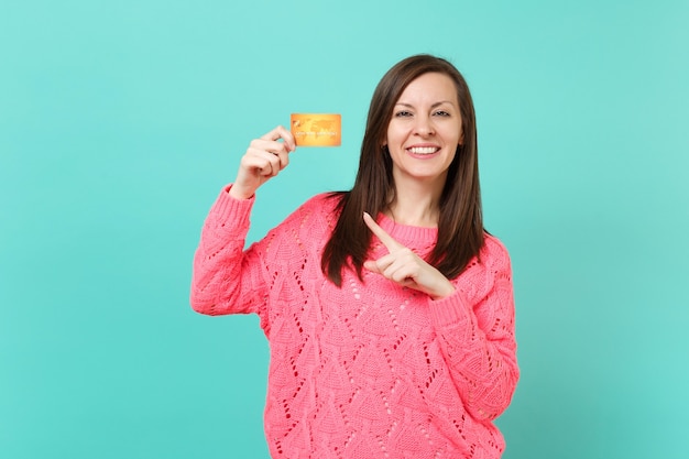 Aantrekkelijke jonge vrouw in gebreide roze trui wijzende wijsvinger op creditcard in de hand geïsoleerd op blauwe turquoise muur achtergrond studio portret. Mensen levensstijl concept. Bespotten kopie ruimte.