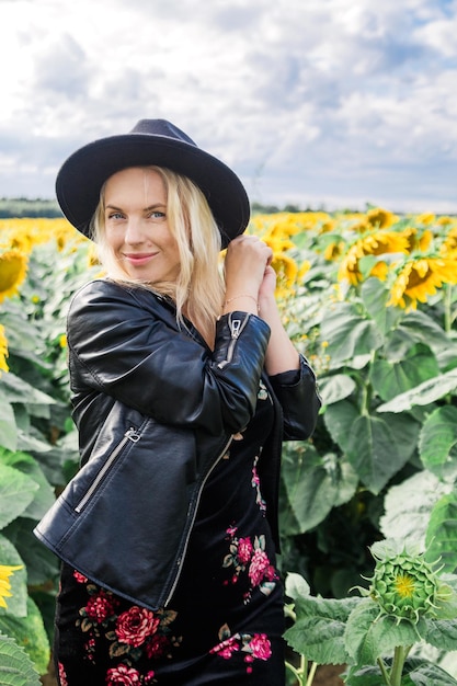 Aantrekkelijke jonge vrouw in jurk zwart leren jas en hoed poseert in het veld met zonnebloemen