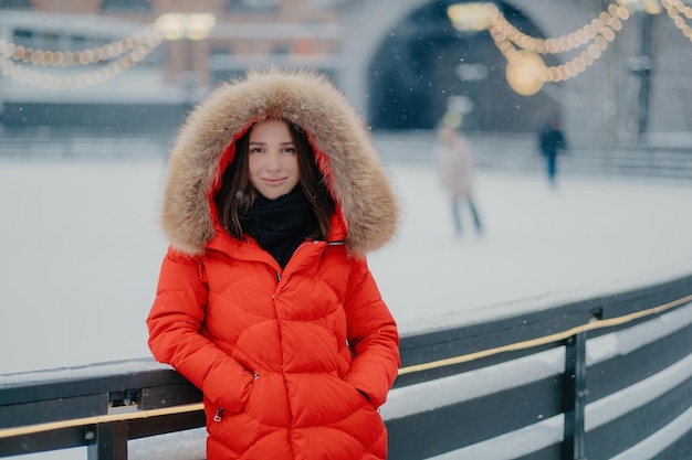 Aantrekkelijke jonge vrouw in rode warme jas staat in de buurt van ijsbaan gaan schaatsen houdt handen in zakken kijkt met zachte glimlach direct naar camera besteedt weekend actief Wintertijd concept