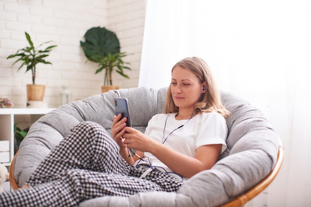 Aantrekkelijke jonge vrouw lwith oortelefoons en smartphonezitting op leunstoelhuis.