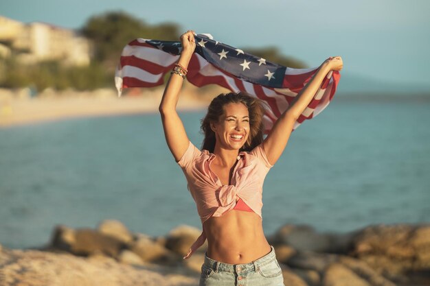 Aantrekkelijke jonge vrouw met Amerikaanse nationale vlag die plezier heeft en een ontspannende dag op het strand heeft.