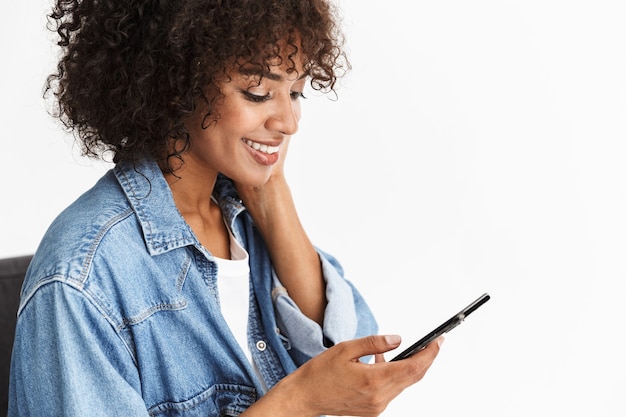 Aantrekkelijke jonge vrouw met denim zittend in een stoel geïsoleerd over een witte muur, met behulp van mobiele telefoon