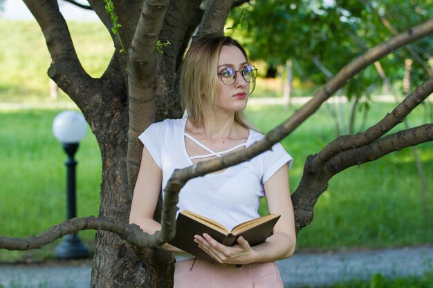 Aantrekkelijke jonge vrouw met een boek in de handen in het park leunend op een boomtak, horizontaal