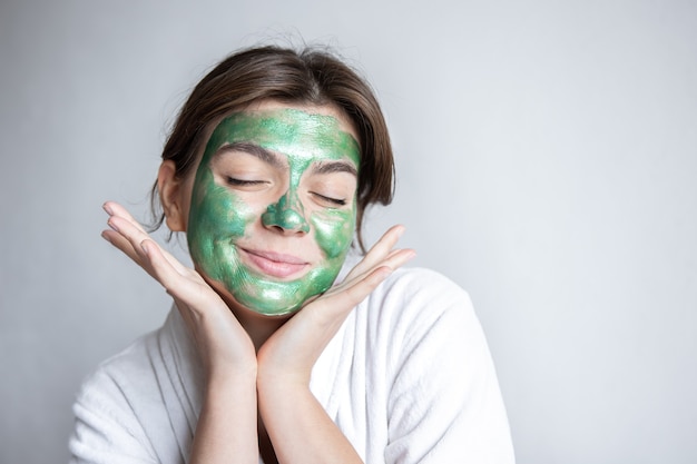 Aantrekkelijke jonge vrouw met een groen cosmetisch masker op haar gezicht en in een wit gewaad op een grijze achtergrond, het concept van spa-behandelingen thuis, kopieer ruimte.