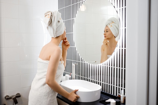 Aantrekkelijke jonge vrouw met een handdoek kijkt naar een spiegel die in de badkamer staat na het douchen Schoonheids- en huidverzorgingsconcept