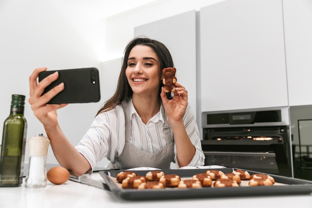 Aantrekkelijke jonge vrouw smakelijke koekjes koken op een dienblad terwijl je in de keuken staat, een selfie te nemen