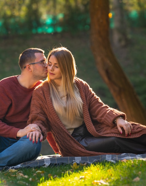 Aantrekkelijke jonge vrouw wordt gekust door haar vriendje op een romantische avonddatum in een park terwijl ze ontspannen op een kleed op het gras