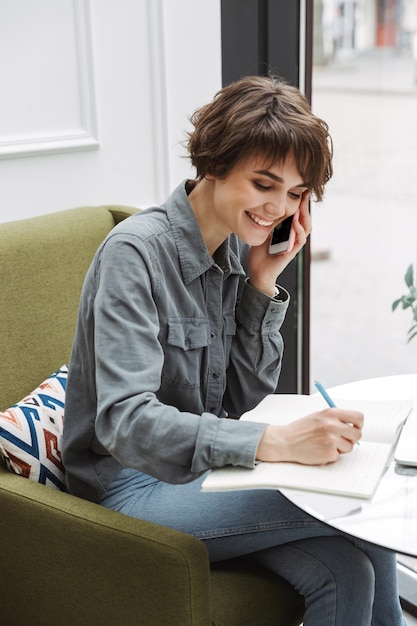 Aantrekkelijke jonge vrouw zittend aan de cafétafel binnenshuis, werken met papierwerk, praten op mobiele telefoon