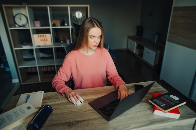 Foto aantrekkelijke jonge vrouwelijke student zit thuis aan haar bureau met boeken en notitieboekjes en gebruikt een laptop