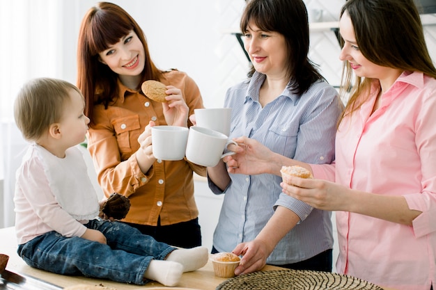 Aantrekkelijke jonge vrouwen, vrouw van middelbare leeftijd en kleine schattige dochter koken op keuken. Liefdevolle familie die hun muffins in de keuken eet en koffie drinkt