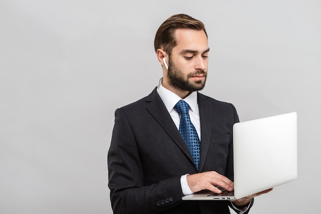 Aantrekkelijke jonge zakenman die een pak draagt dat geïsoleerd staat over een grijze muur, met behulp van een laptopcomputer