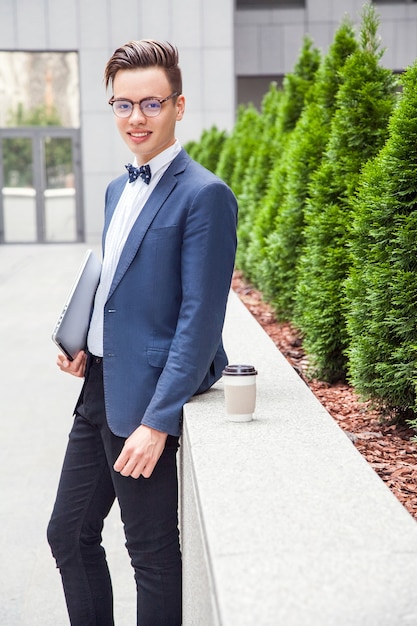 Aantrekkelijke jonge zakenman met laptop en koffie in handen op kantoorgebouw achtergrond. laptop en koffie vasthouden en naar de camera kijken met een brede glimlach.