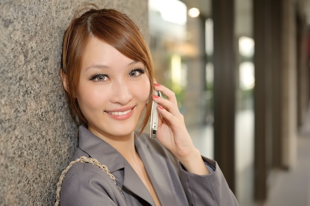Aantrekkelijke jonge zakenvrouw met mobiele telefoon tegen muur en op zoek naar jou, halve lengte close-up portret buiten moderne gebouwen.