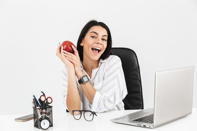 Aantrekkelijke jonge zakenvrouw zittend aan het bureau geïsoleerd over witte muur, met rode appel