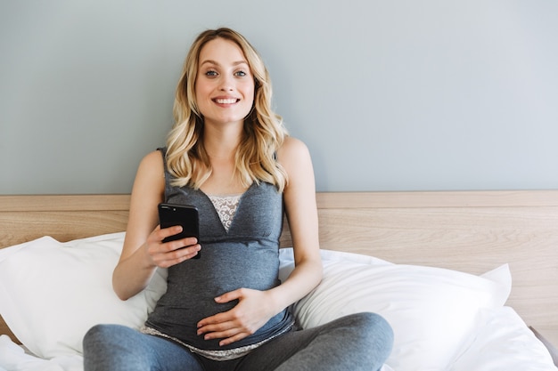 Aantrekkelijke jonge zwangere blonde vrouw ontspannen op bed thuis, met behulp van mobiele telefoon