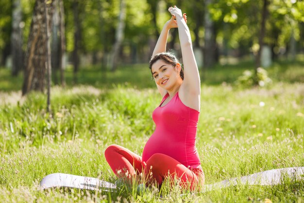 Aantrekkelijke jonge zwangere vrouw die yogaoefeningen in openlucht in het park doet. Verwacht dat een vrouw een gezonde en actieve levensstijl heeft.