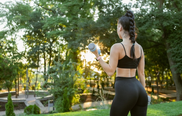 Aantrekkelijke Kaukasische fit brunette vrouw training biceps spieren met halters in haar handen in het park bij zonsopgang. Achteraanzicht