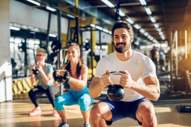 Aantrekkelijke knappe bebaarde man kettlebell te houden en squats te doen in een fitnessgroep met twee meisjes in de moderne sportschool.