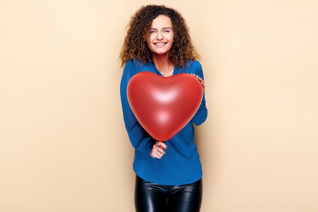 Aantrekkelijke krullende haarvrouw die de rode ballon van de hartvorm houdt