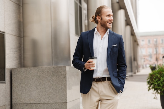 Aantrekkelijke lachende jonge blonde harige man in formele kleding praten via oortelefoons, afhaalmaaltijden koffiekopje houden terwijl hij buiten op een straat in de stad
