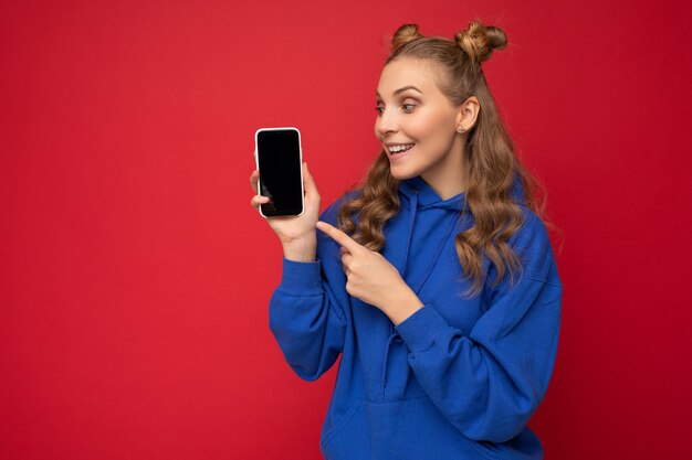 Aantrekkelijke lachende jonge blonde vrouw dragen stijlvolle blauwe hoodie geïsoleerd op rode achtergrond met kopie ruimte smartphone weergegeven: telefoon in de hand met leeg scherm wijzend op gadjet looki