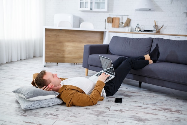 Aantrekkelijke lachende jonge man in vrijetijdskleding zittend op de vloer op de bank in de woonkamer met behulp van laptopcomputer