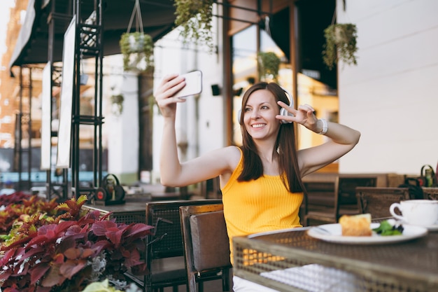 Aantrekkelijke lachende vrouw in openlucht straat coffeeshop café aan tafel zitten, muziek luisteren in koptelefoon, selfie geschoten op mobiele telefoon doen, ontspannen in restaurant vrije tijd